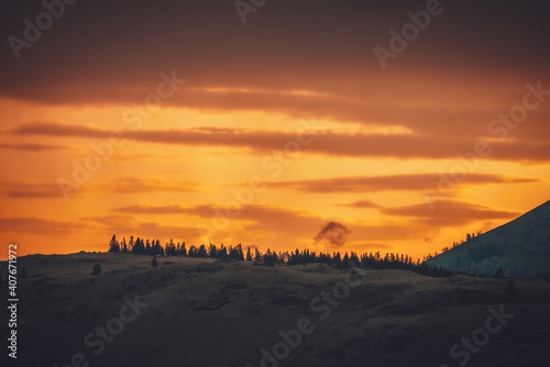 Atmospheric landscape with forest silhouette on silhouette of mountain on background of vivid orange dawn sky. Colorful nature scenery with sunset or sunrise of illuminating color. Sundown paysage.