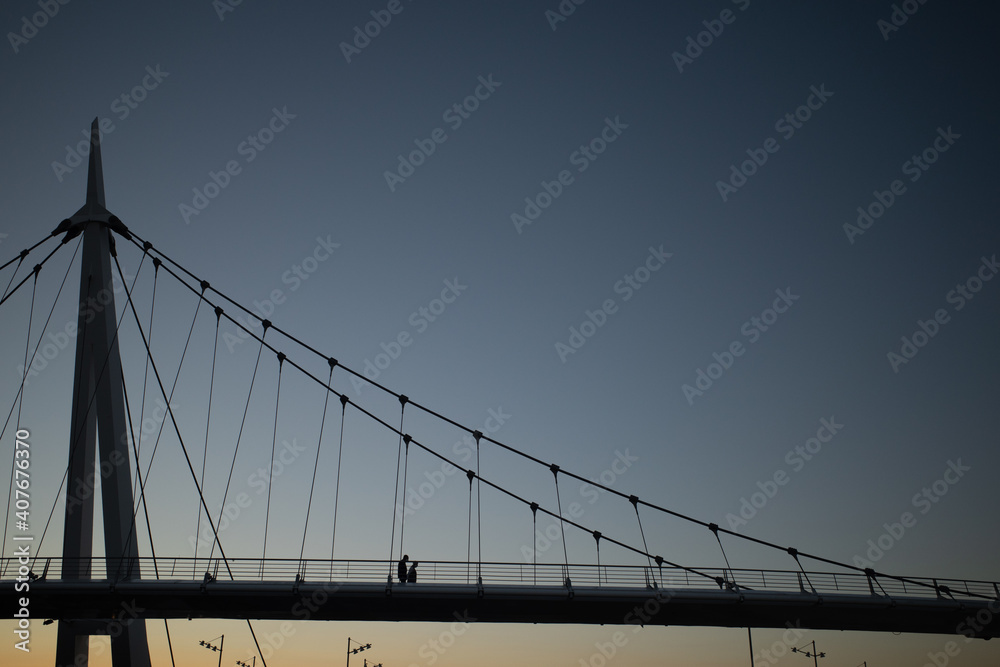Siluetas de pareja caminando sobre puente colgante moderno al atardecer Siluetas de pareja caminando sobre puente colgante moderno al atardecer