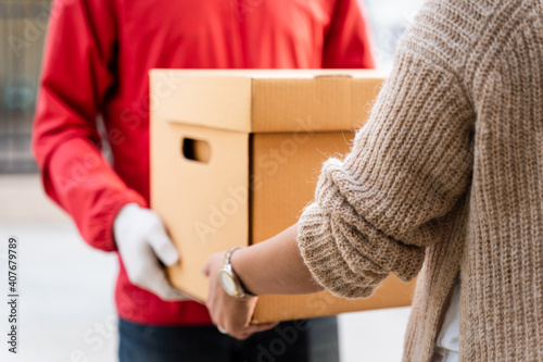 An Asian deliver man in red uniform handing parcel to a female costumer in front of the house. A postman and express delivery service deliver parcel during covid19 pandemic.