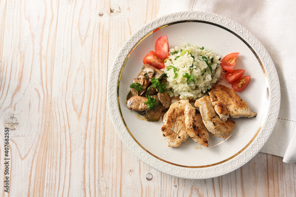 Fried chicken slices with cauliflower rice, mushroom ragout and tomatoes, on a light wooden table, healthy slimming with low carb diet, copy space, high angle view from above
