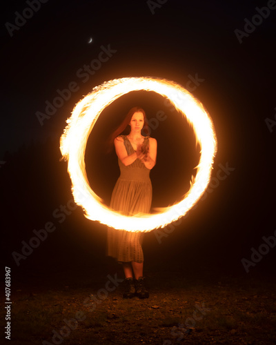 Graceful elegant red haired female fireshow performer making fire dance with two metal fans lit with flames on night mountains meadow photo