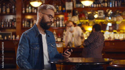 Bearded man texting on smartphone sitting at modern pub