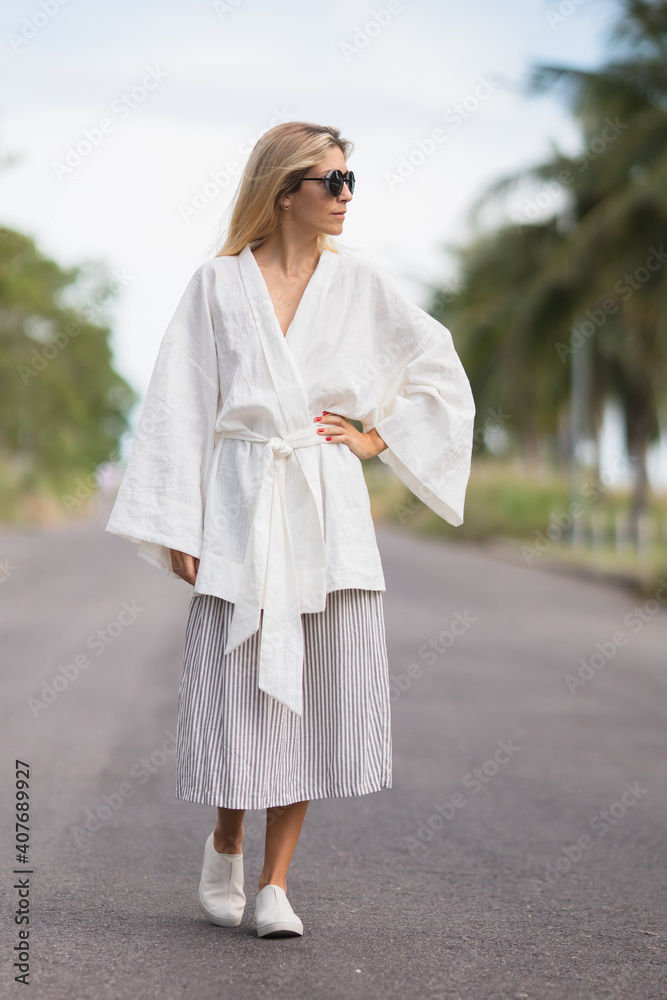 Woman is walking outside on the street wearing white linen kimono robe, striped silk skirt and white leather shoes