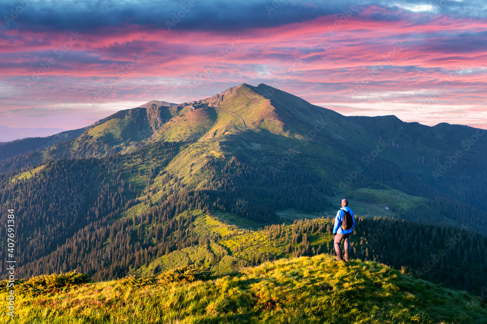 A tourist on the edge of a mountain covered with a lush grass. Purple sunset sky and hight mountains peaks on background. Landscape photography