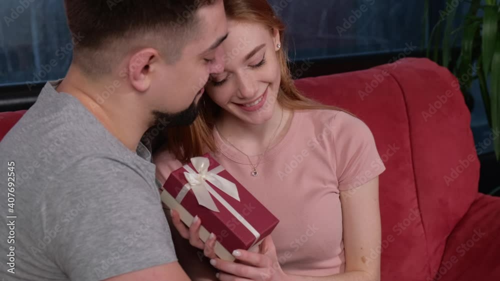 Ginger lady with freckles is receiving a present from her lover on valentines day embracing and smile on a couch Ginger lady with freckles is receiving a present from her lover on valentines day embracing and smile on a couch