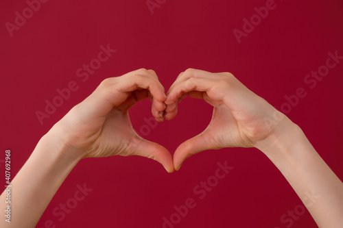 Female hands showing heart sign on red background in studio. Fingers illustrating gesture of love on burgundy backdrop. Valentine theme expressed in symbol.