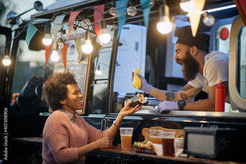young beardy caucasian employee in fast food laughing, adding a  mustard in a sandwich to a female afro-american customer