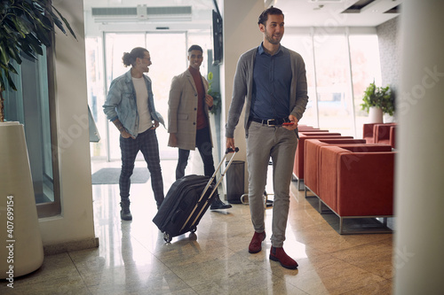 young businessmen entering hotel, caring luggage, suitcase on wheels, talking, smiling, laughing