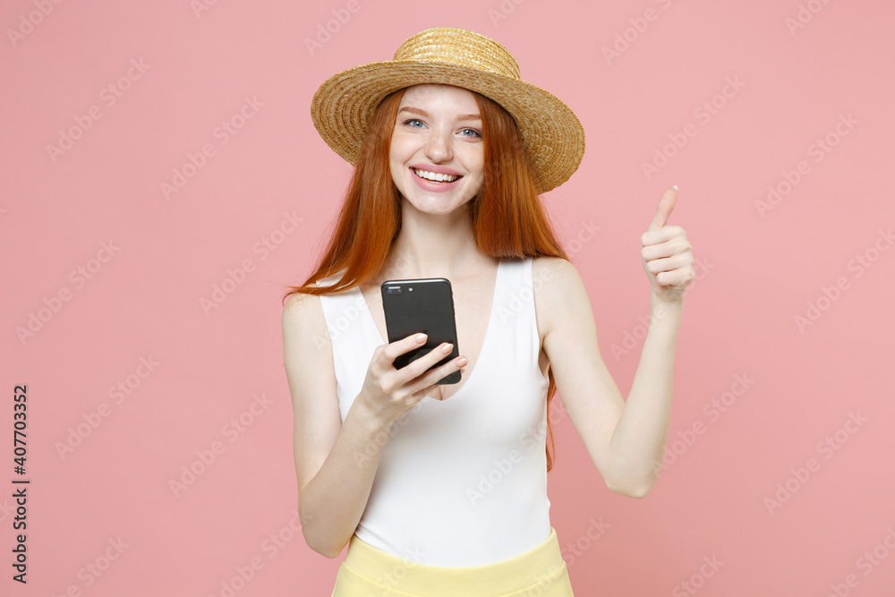 Young smiling gladden redhead woman 20s ginger long hair in straw hat summer clothes hold in hand mobile cell phone showing thumbs up like gesture isolated on pastel pink background studio portrait Young smiling gladden redhead woman 20s ginger long hair in straw hat summer clothes hold in hand mobile cell phone showing thumbs up like gesture isolated on pastel pink background studio portrait