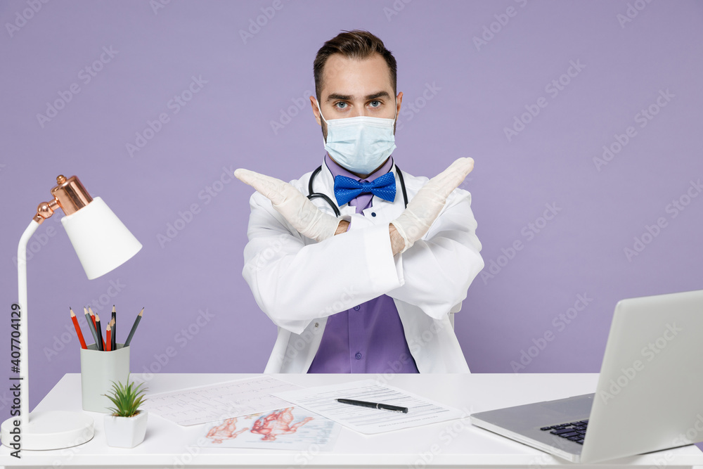Male doctor man in white medical suit face mask gloves from coronavirus covid-19 sit at desk in clinic office point fingers crossed hands on copy space isolated on violet background studio portrait. Male doctor man in white medical suit face mask gloves from coronavirus covid-19 sit at desk in clinic office point fingers crossed hands on copy space isolated on violet background studio portrait.