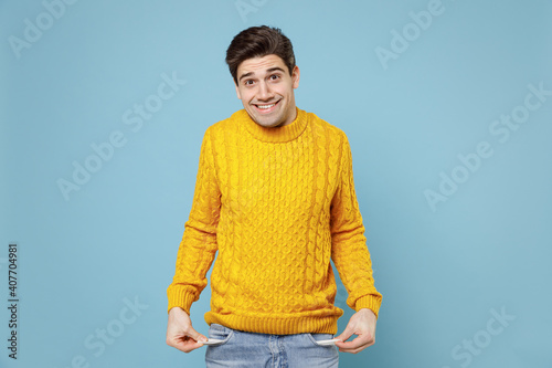 Young caucasian penniless cheerful poor student man 20s in casual knitted yellow fashionable sweater looking camera showing empty pants jeans pockets isolated on blue color background studio portrait.