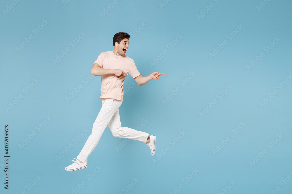 Full length of young surprised cheerful smiling student man 20s in beige t-shirt white pants point index finger aside on copy space workspace area jump run isolated on blue background studio portrait.