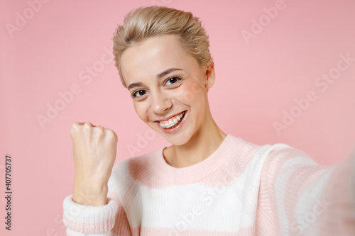 Close up of young smiling woman short haircut nude makeup dental braces in sweater do selfie shot on mobile phone show winner gesture clenching fist isolated on pastel pink background studio portrait.