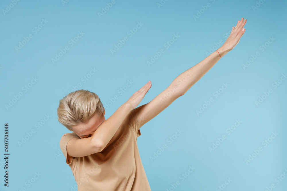 Young blonde woman short haircut in casual basic beige t-shirt doing dab hip hop dance hands move gesture youth sign hiding covering face celebrating isolated on blue color background studio portrait. Young blonde woman short haircut in casual basic beige t-shirt doing dab hip hop dance hands move gesture youth sign hiding covering face celebrating isolated on blue color background studio portrait.