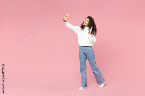 Full length young happy smiling african american woman 20s in white knitted sweater jeans do selfie shot on mobile phone show victory v-sign gesture isolated on pastel pink background studio portrait. © ViDi Studio