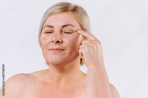 Caucasian blond woman applying cream mask with closed eyes. Beauty concept on white background.