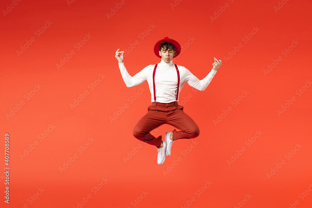 Full length of young spanish latinos relaxed man 20s in hat white shirt trousers, suspenders jump high meditating do yoga gesture closed eyes levitating isolated on red background studio portrait.