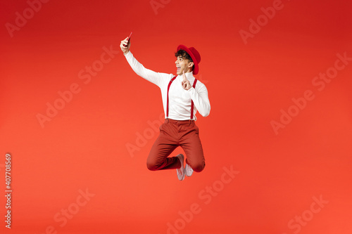 Full length of young spanish latinos smiling stylish fashionable man 20s in hat white shirt trousers, suspenders walk go doing selfie shot on mobile phone isolated on red background studio portrait.