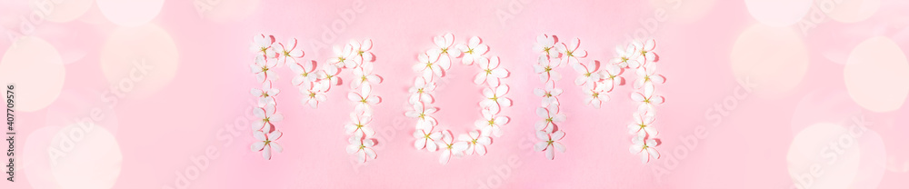 Fototapeta premium Wide festive banner with word MOM laid out from flowers of white apple tree on pink backdrop with beautiful bokeh. Holiday Mother's day floral concept. Top view, flat lay.