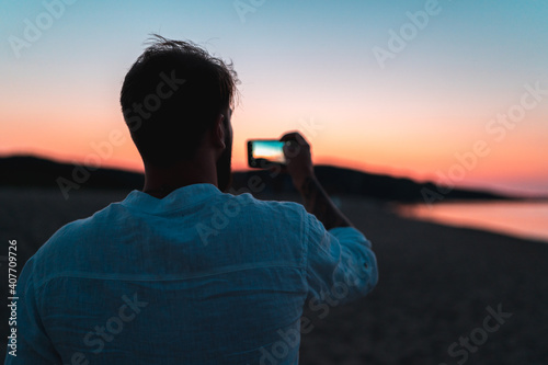 Back view of young man taking photo of colorful sunset over the sea coast using his smartphone