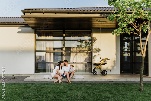 A young beautiful family with two little sons hug and kiss in the yard of their own house. Young mom and dad with two young sons hug and kiss in the yard of their own house.