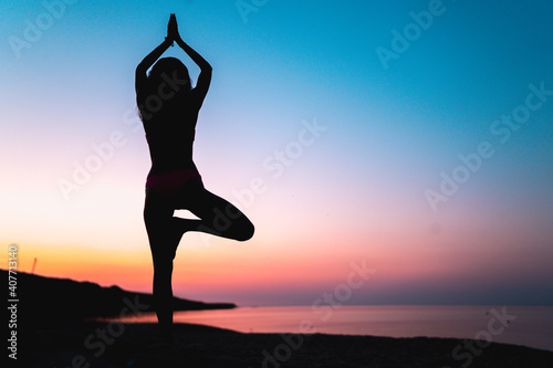 Silhouette Of Fit Woman in Vrikshasana Tree Yoga Pose on the back with colorful sunset and reflecting sea surface