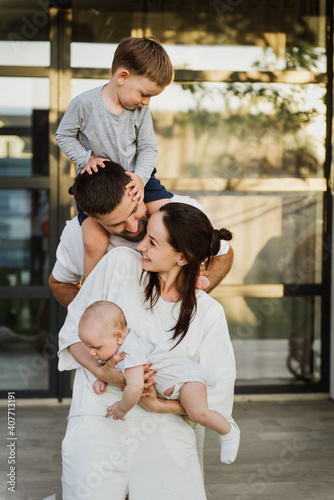 A young beautiful family with two little sons hug and kiss in the yard of their own house. Young mom and dad with two young sons hug and kiss in the yard of their own house.