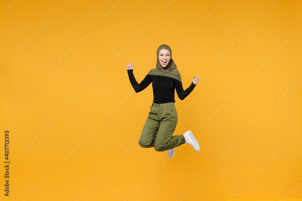 Full length happy young arabian muslim woman in hijab black green clothes jumping clenching fists like winner say yes isolated on yellow background studio portrait. People religious lifestyle concept.