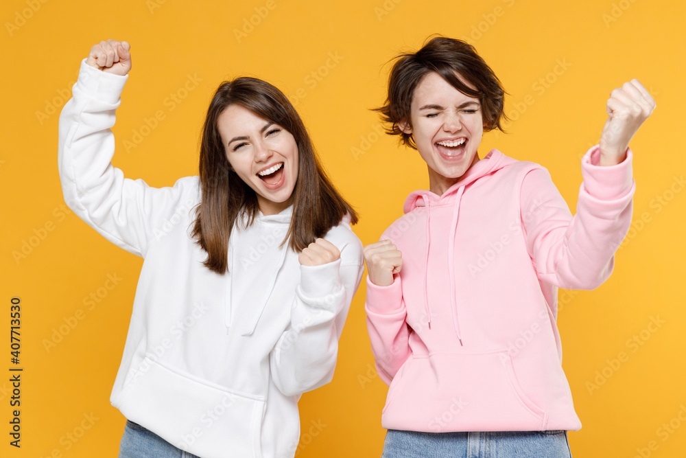Happy joyful two young women friends 20s wearing casual white pink hoodies doing winner gesture celebrating clenching fists say yes isolated on bright yellow color wall background studio portrait. Happy joyful two young women friends 20s wearing casual white pink hoodies doing winner gesture celebrating clenching fists say yes isolated on bright yellow color wall background studio portrait.