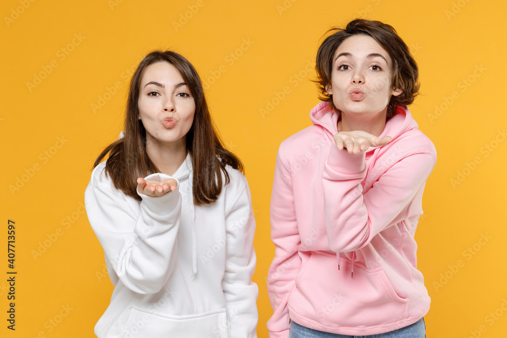 Pretty charming beautiful two young women friends 20s wearing casual white pink hoodies standing blowing sending air kiss looking camera isolated on bright yellow color background studio portrait. Pretty charming beautiful two young women friends 20s wearing casual white pink hoodies standing blowing sending air kiss looking camera isolated on bright yellow color background studio portrait.