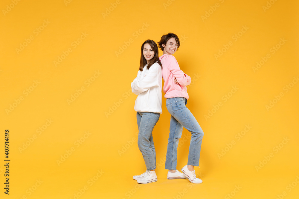 Full length side view of cheerful two young women friends 20s wearing casual white pink hoodies standing back to back holding hands crossed isolated on bright yellow color background studio portrait. Full length side view of cheerful two young women friends 20s wearing casual white pink hoodies standing back to back holding hands crossed isolated on bright yellow color background studio portrait.