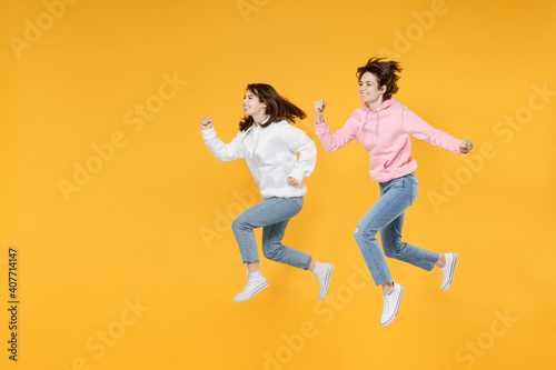 Full length side view of cheerful two young women friends 20s wearing basic white pink hoodies jumping running clenching fists like winner isolated on bright yellow color background studio portrait.