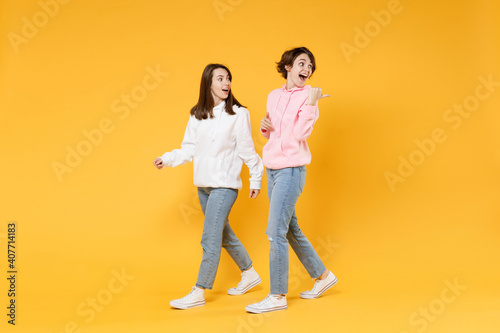 Full length side view of excited shocked two young women friends 20s wearing casual white pink hoodies walking going pointing thumb aside isolated on bright yellow color background studio portrait.