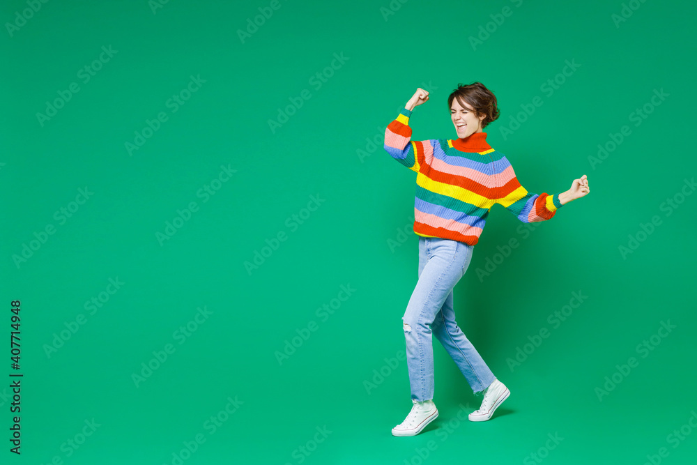 Full length side view of happy joyful young brunette woman 20s in colorful sweater doing winner gesture celebrating clenching fists say yes isolated on bright green color background studio portrait. Full length side view of happy joyful young brunette woman 20s in colorful sweater doing winner gesture celebrating clenching fists say yes isolated on bright green color background studio portrait.
