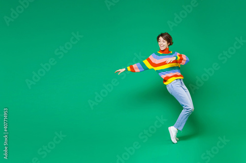 Full length side view of cheerful funny young brunette woman 20s in basic casual colorful sweater dancing standing on toes spreading hands isolated on bright green color background studio portrait.