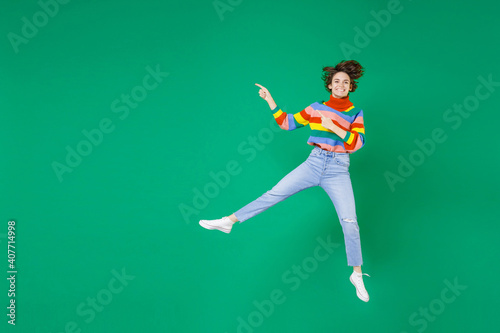 Full length of smiling cheerful funny young brunette woman 20s in colorful sweater jumping pointing index fingers aside on mock up copy space isolated on bright green color background studio portrait.