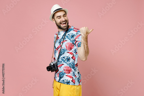 Side view of funny young traveler tourist man in summer clothes hat with photo camera point thumb aside isolated on pink background. Passenger traveling abroad on weekends. Air flight journey concept.
