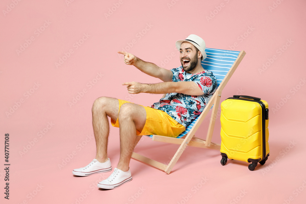 Full length excited young traveler tourist man in summer clothes hat sit on deck chair point index fingers aside isolated on pink background. Passenger travel on weekend. Air flight journey concept. - obrazy, fototapety, plakaty 