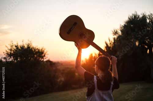 Ragazza di spalle solleva la chitarra al cielo durante il momento del tramonto in contesto naturale photo