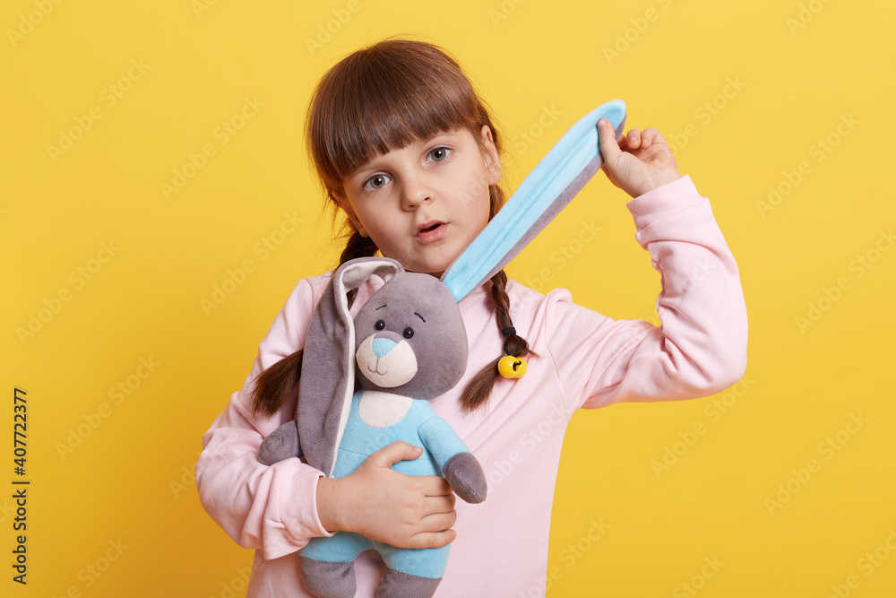 Adorable little girl with toy rabbit isolated over yellow background, child pulling bunny's ear and loos at camera with astonished and serious facial expression. Adorable little girl with toy rabbit isolated over yellow background, child pulling bunny's ear and loos at camera with astonished and serious facial expression.