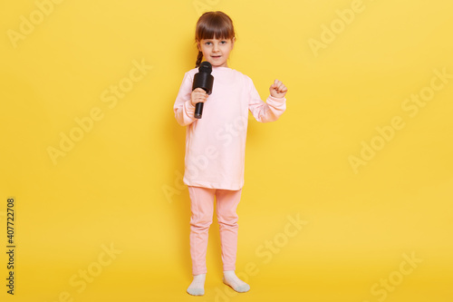 Close up photo of cool attractive lady kid having fun, standing isolated over yellow background, adorable kid with microphone arranging concert, dresses pale pink background. © sementsova321