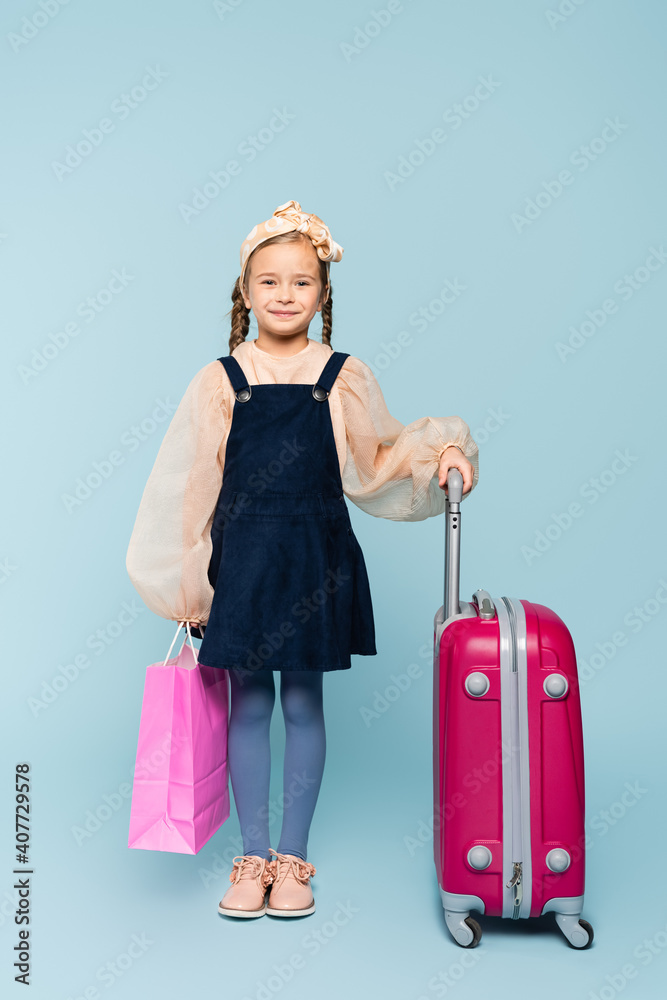 full length of happy little girl holding shopping bag and standing with baggage on blue full length of happy little girl holding shopping bag and standing with baggage on blue