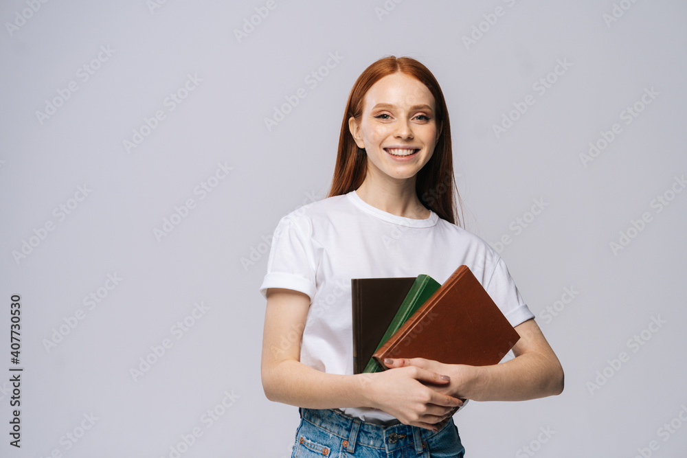 Cheerful young woman college student holding books and looking at camera on isolated gray background. Pretty redhead lady model wearing casual fashion clothes emotionally showing facial expressions. Cheerful young woman college student holding books and looking at camera on isolated gray background. Pretty redhead lady model wearing casual fashion clothes emotionally showing facial expressions.