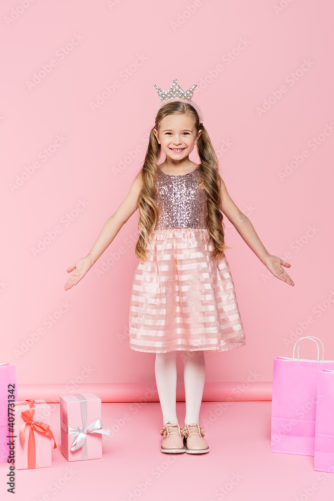 full length of happy little girl in crown standing near presents and shopping bags on pink