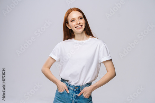 Cheerful young woman wearing T-shirt and denim pants holds hands in pockets on isolated white background. Pretty lady model with red hair emotionally showing facial expressions in studio, copyspace.