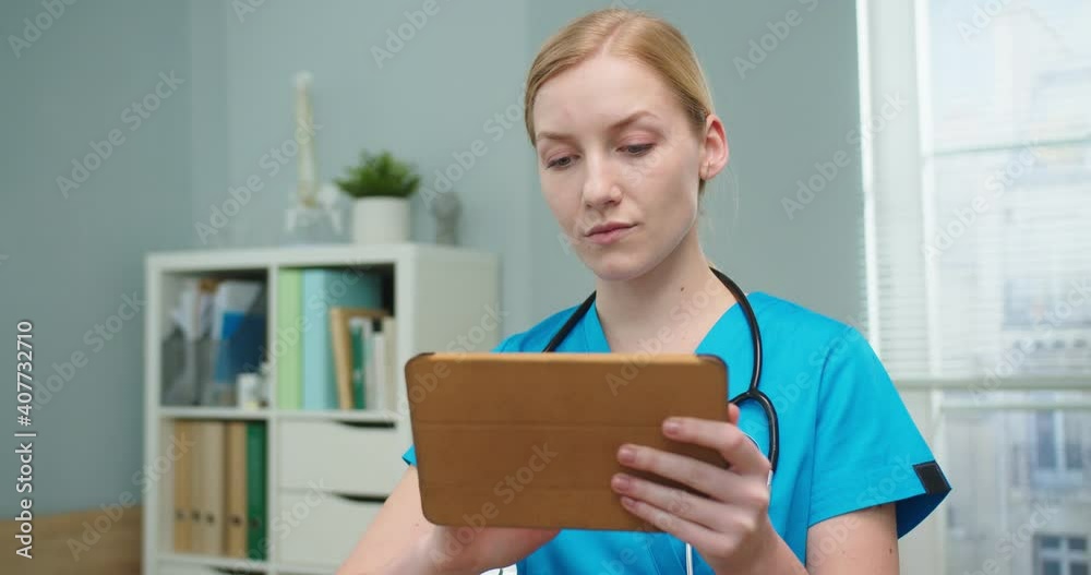 Portrait of charming female nurse wearing blue medical uniform using tablet in doctor's office. Young Caucasian woman typing on gadget and looking at camera. Technology, healthcare concept. Portrait of charming female nurse wearing blue medical uniform using tablet in doctor's office. Young Caucasian woman typing on gadget and looking at camera. Technology, healthcare concept.
