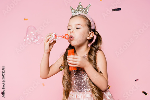 little girl in crown blowing soap bubbles near falling confetti on pink