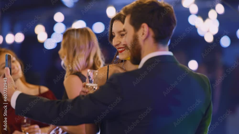 Caucasian Beautiful Nice Couple Recording Selfie Video on Smartphone Camera Celebrating Night Holiday Corporate Party with Business Friends. Caucasian Beautiful Nice Couple Recording Selfie Video on Smartphone Camera Celebrating Night Holiday Corporate Party with Business Friends.