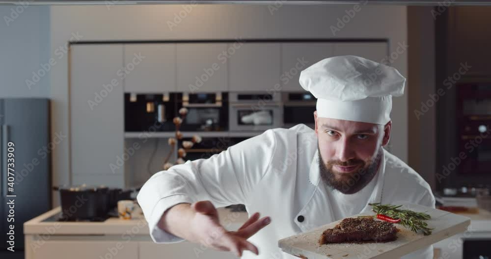 Young handsome chef in white uniform holding wooden board with ready beef steak Young handsome chef in white uniform holding wooden board with ready beef steak