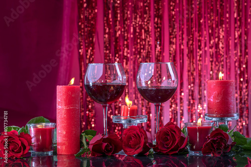 Lit red candles in transparent candlesticks illuminate glasses with wine surrounded by roses on a mirror surface on a red shining bokeh background. Valentine's day or romantic evening invitation